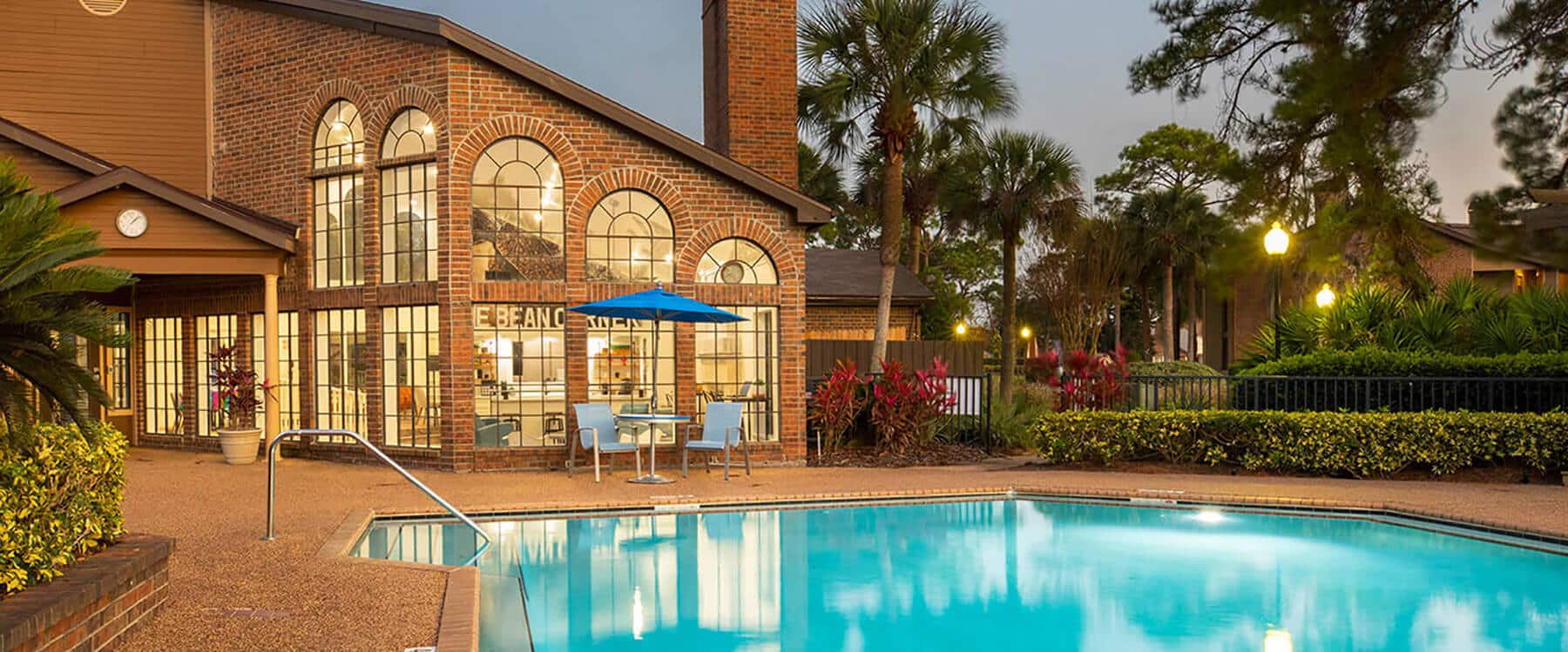 A swimming pool in front of a brick apartment building with large windows, outdoor seating, palm trees, and evening lighting evokes the relaxed elegance of Anatole living.