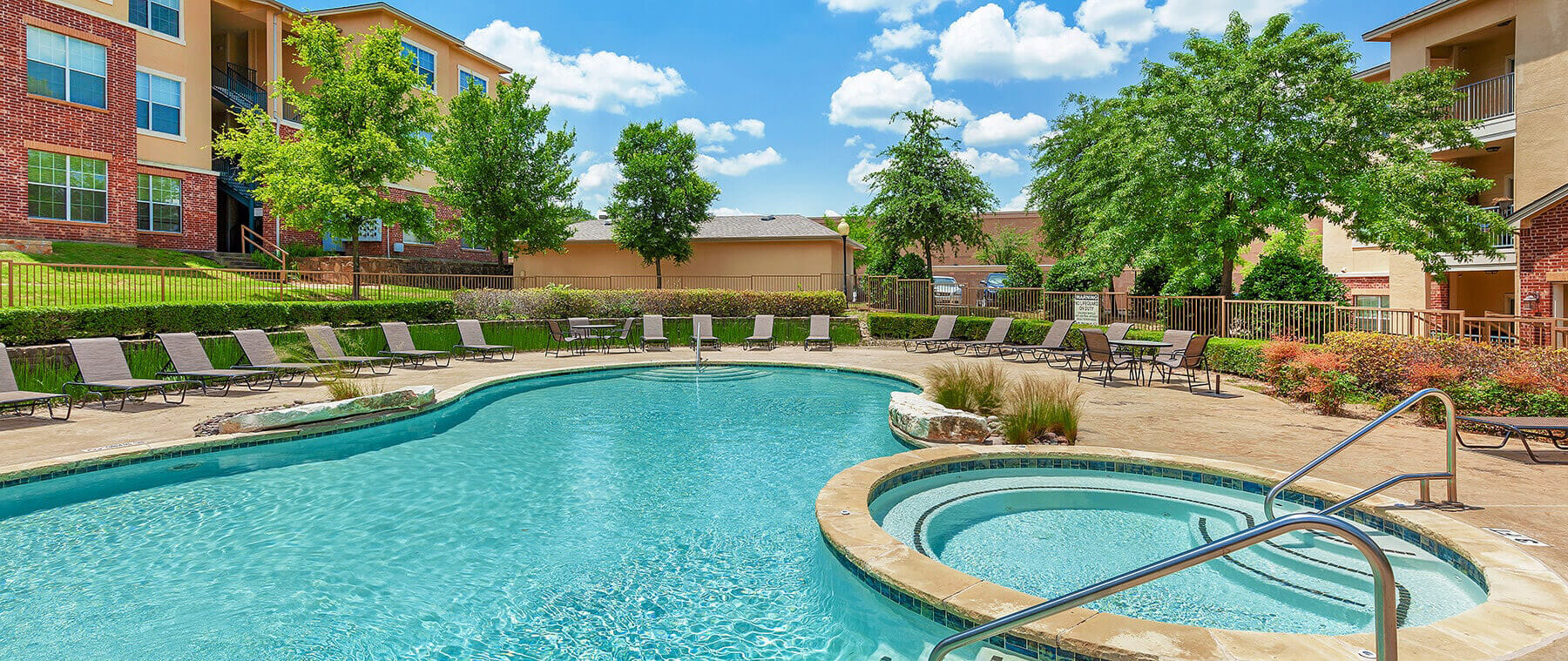 Enjoy the beautiful outdoor swimming pool area at Bella Ruscello, complete with a relaxing hot tub, surrounded by lounge chairs, lush trees, and apartment buildings under a picturesque blue sky with clouds.
