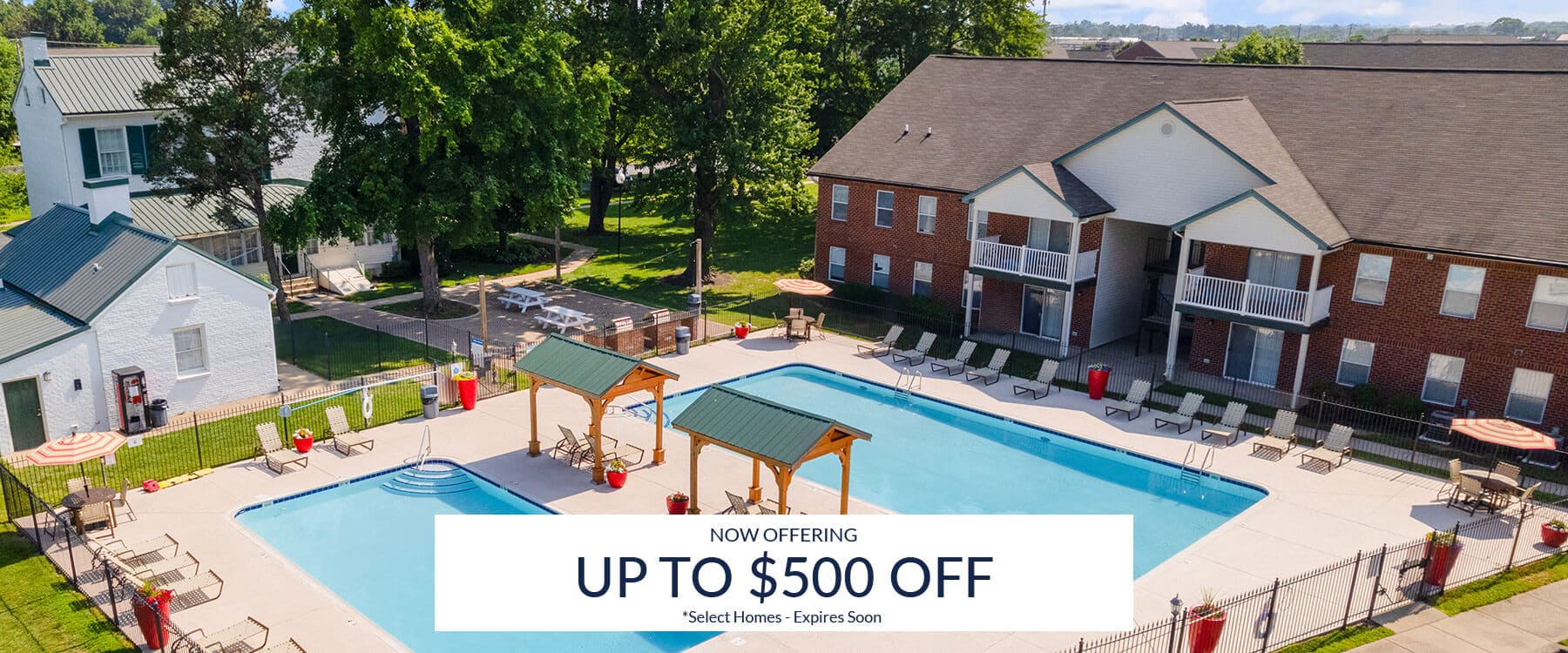 A large outdoor swimming pool with lounge chairs and shaded seating, adjacent to brick apartment buildings; a banner announces up to $500 off select homes.