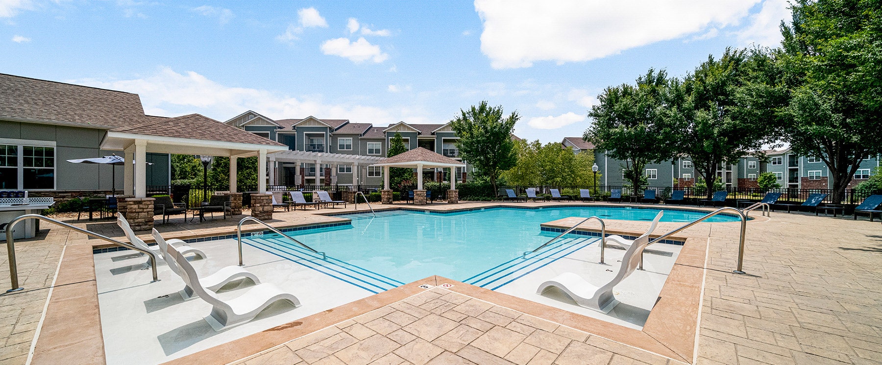 Creekside outdoor swimming pool area with lounge chairs and pergolas, surrounded by apartment buildings and trees on a sunny day.