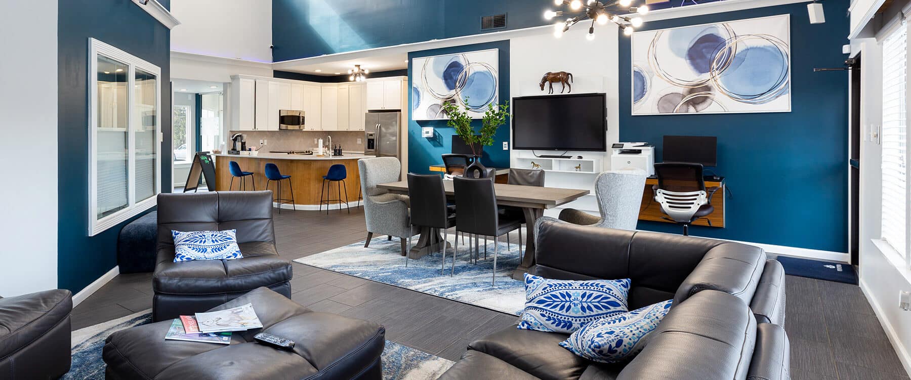 Modern open-concept living and dining area with blue accents, leather sofas, steeplechase-inspired wall art, a dining table, and a kitchen in the background.